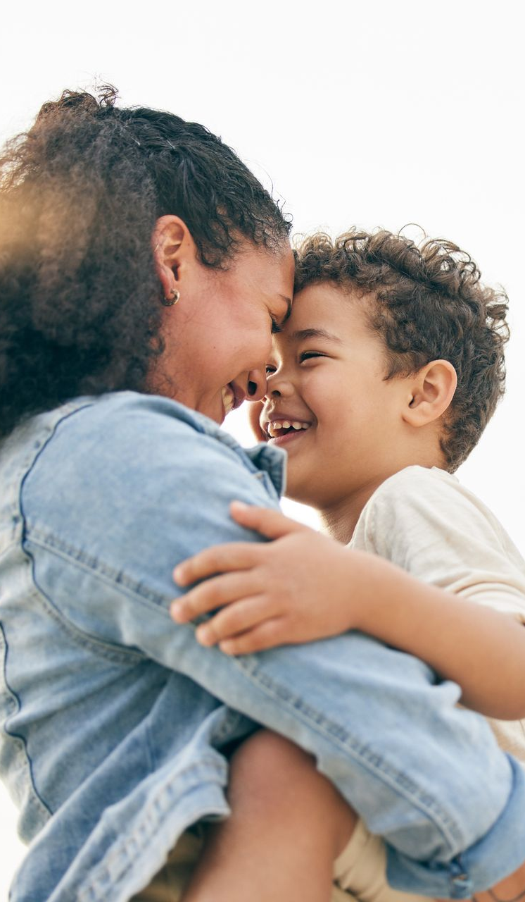 Woman hugs child, both smiling, foreheads touching, outside.