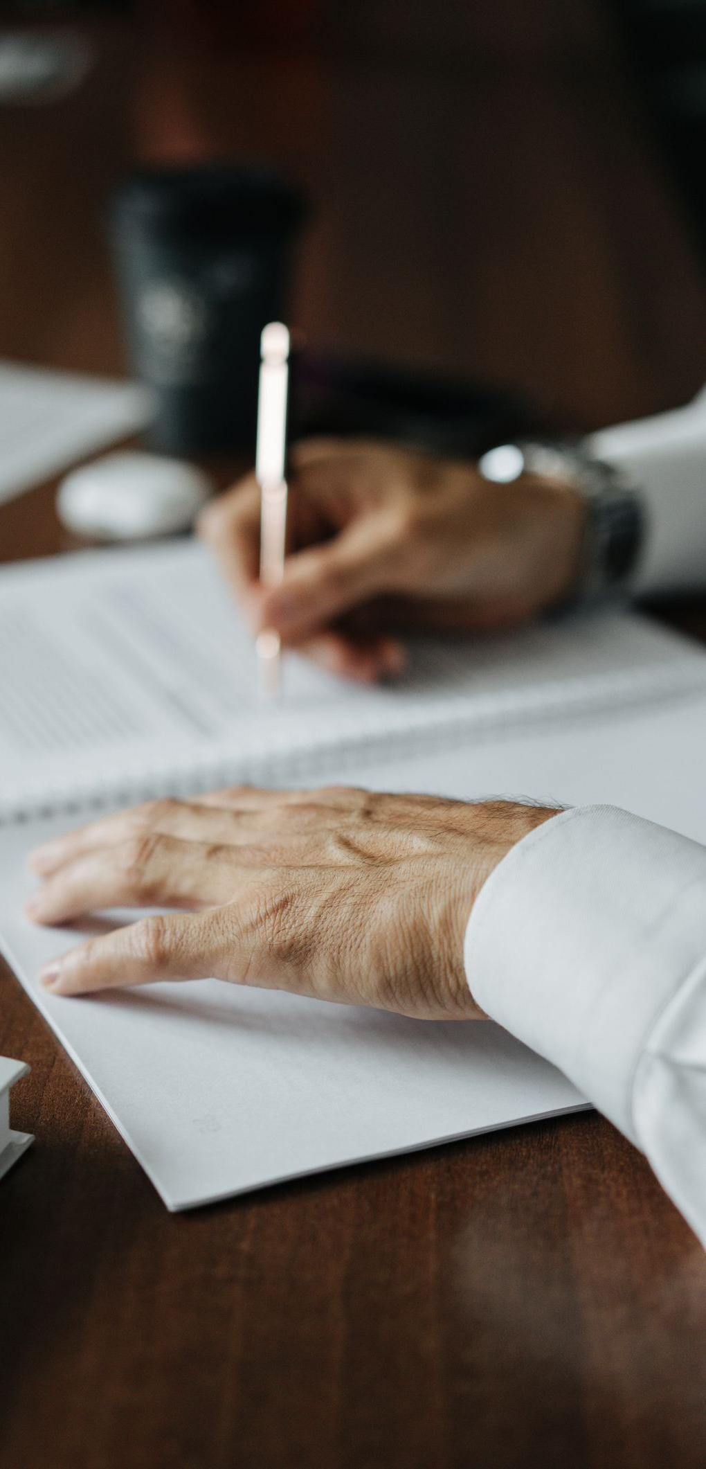 A person's hands writing on a white paper with a pen on a dark wooden table.