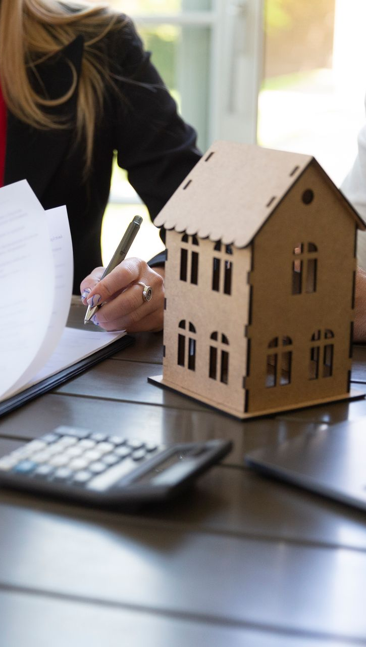 Woman signing papers next to a model house and calculator on a table.