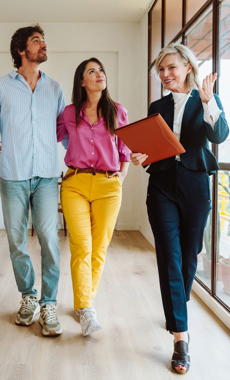 Real estate agent showing a couple a home. Agent gestures, couple looks up. Bright, airy room.