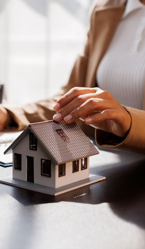 Woman's hand on a miniature house model on a desk. Brown blazer, white top, and natural light.
