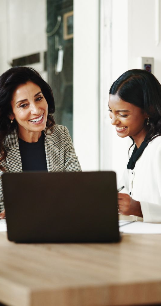Two women smiling, working together at a laptop and documents on a desk.