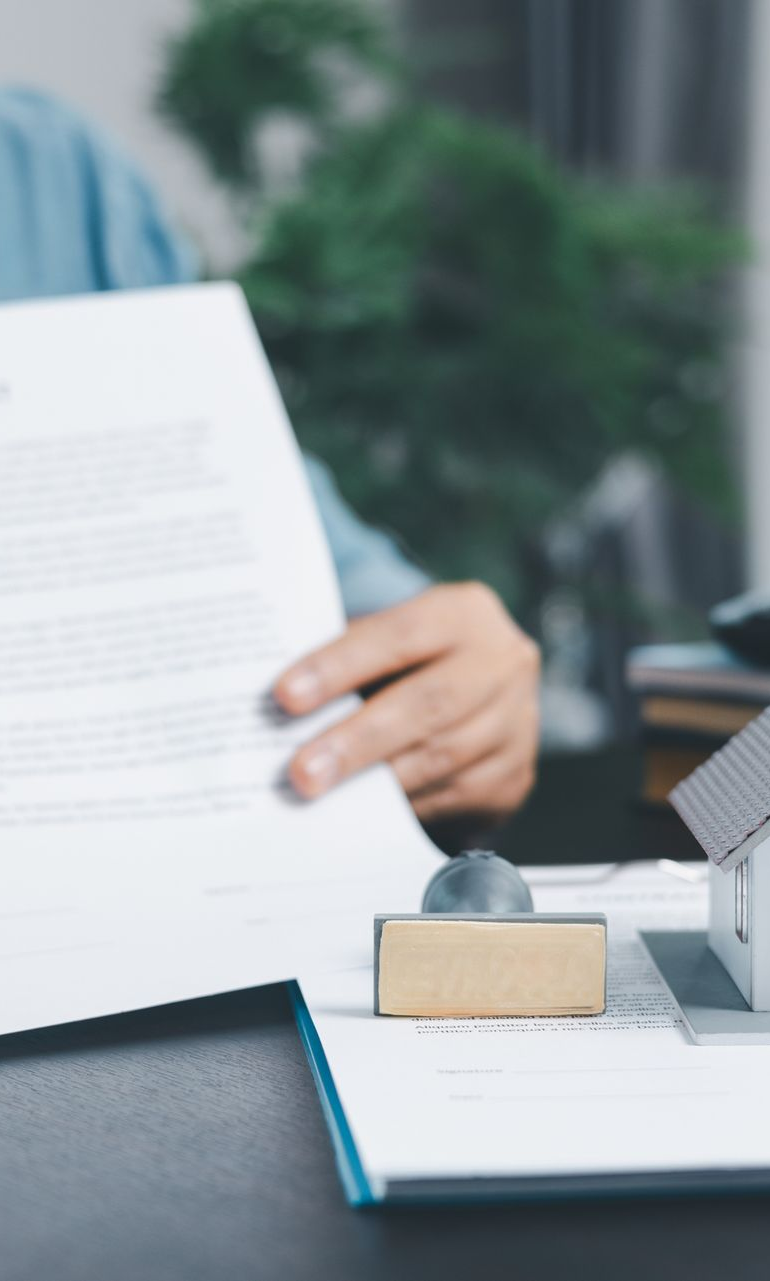 Person holding documents above a desk with a stamp and miniature house.