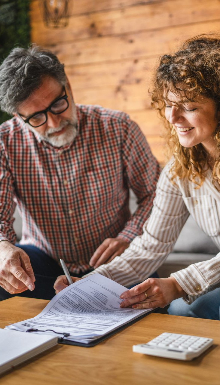 Man with glasses and woman signing documents at a table; calculator visible.