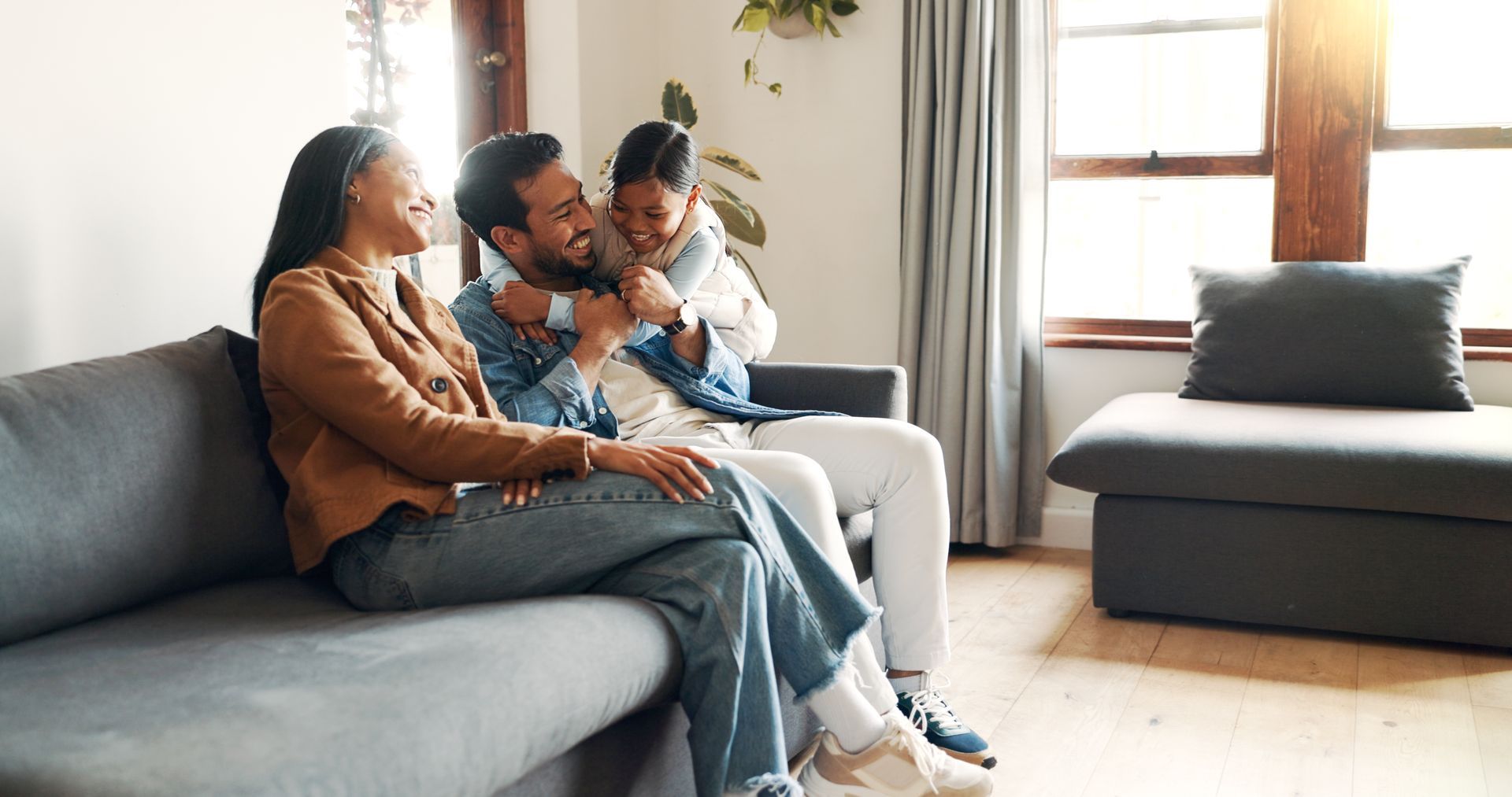 Family of three smiling, embracing on a couch in a sunlit living room.