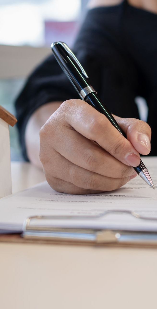 Person signing document with pen, small house model in background.
