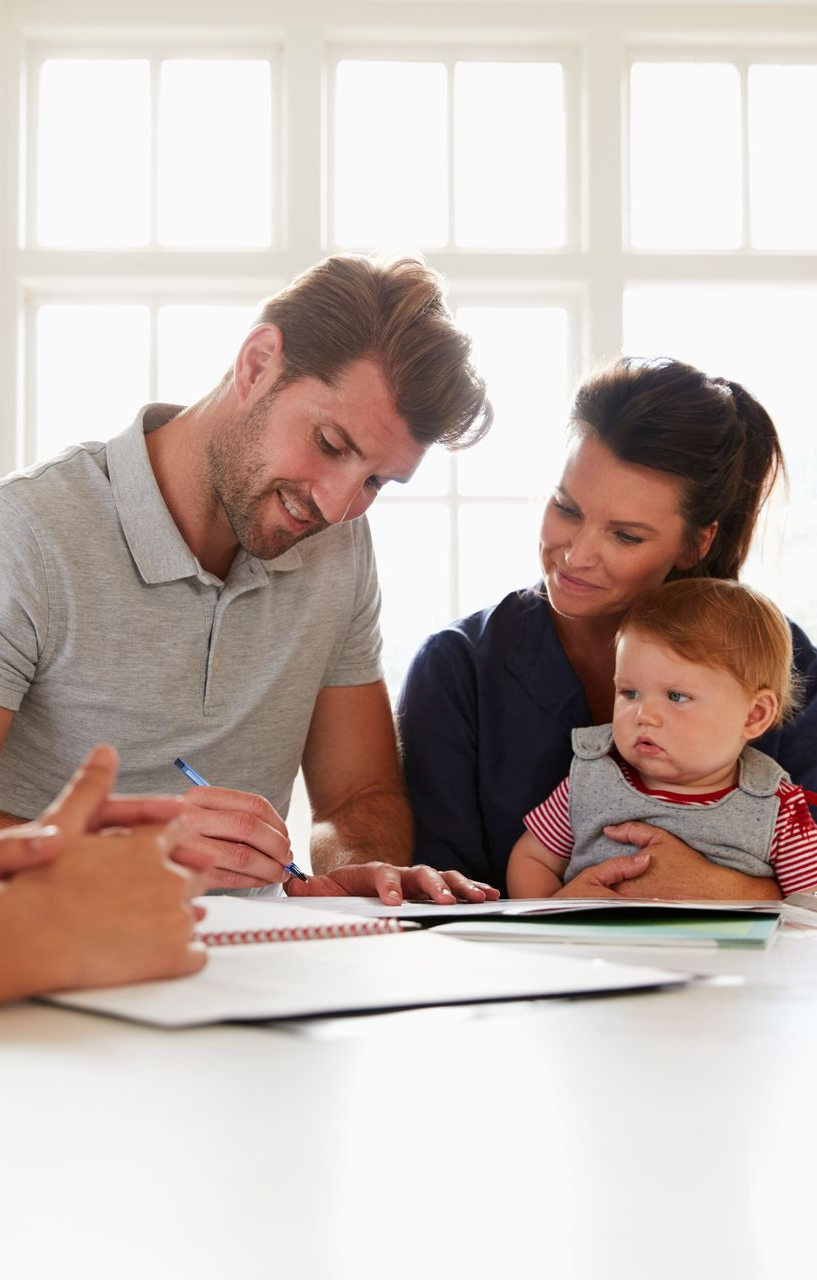 Family sitting at table; man signing document, woman holding baby, bright room.