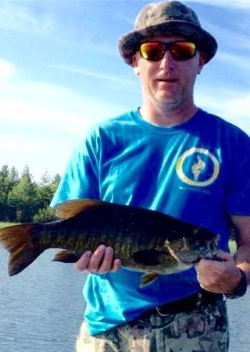 Person holding a fish outdoors by the water, wearing a blue shirt, hat, and sunglasses.