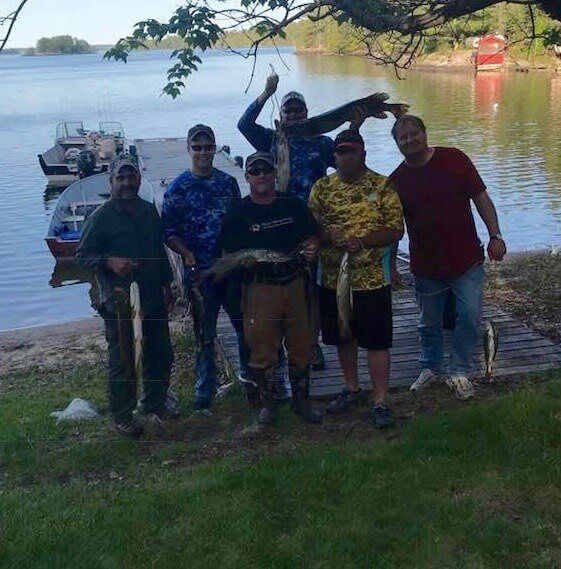 Six people posing on a lakeside dock with a boat and calm water in the background.
