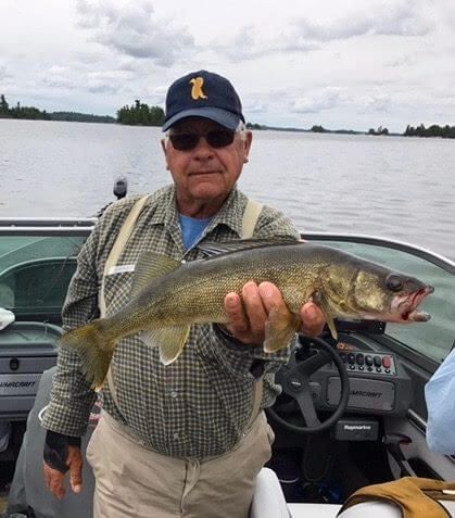 Man holding a large fish on a boat, with a lake and cloudy sky in the background.