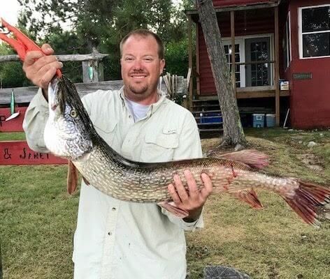 Man holding a large fish by the tail in a grassy outdoor setting near a rustic building.