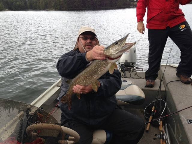 Angler on a boat holds up a large fish on a lake while another person stands nearby.