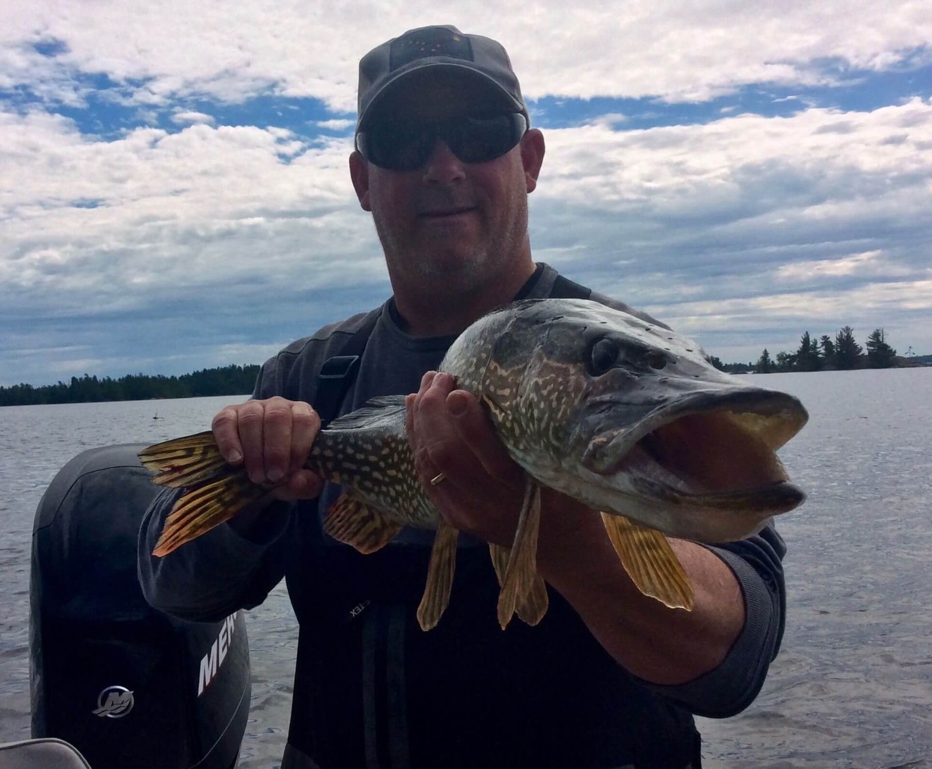 Person holding a large fish on a boat, wearing sunglasses and cap, with open water and cloudy sky behind.