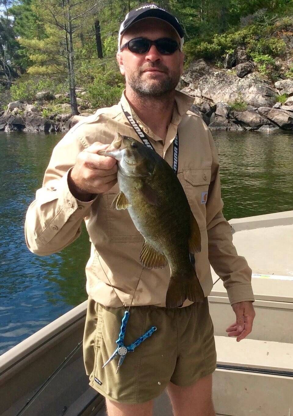 Man on a boat holding a largemouth bass over a calm lake with rocky shoreline behind him