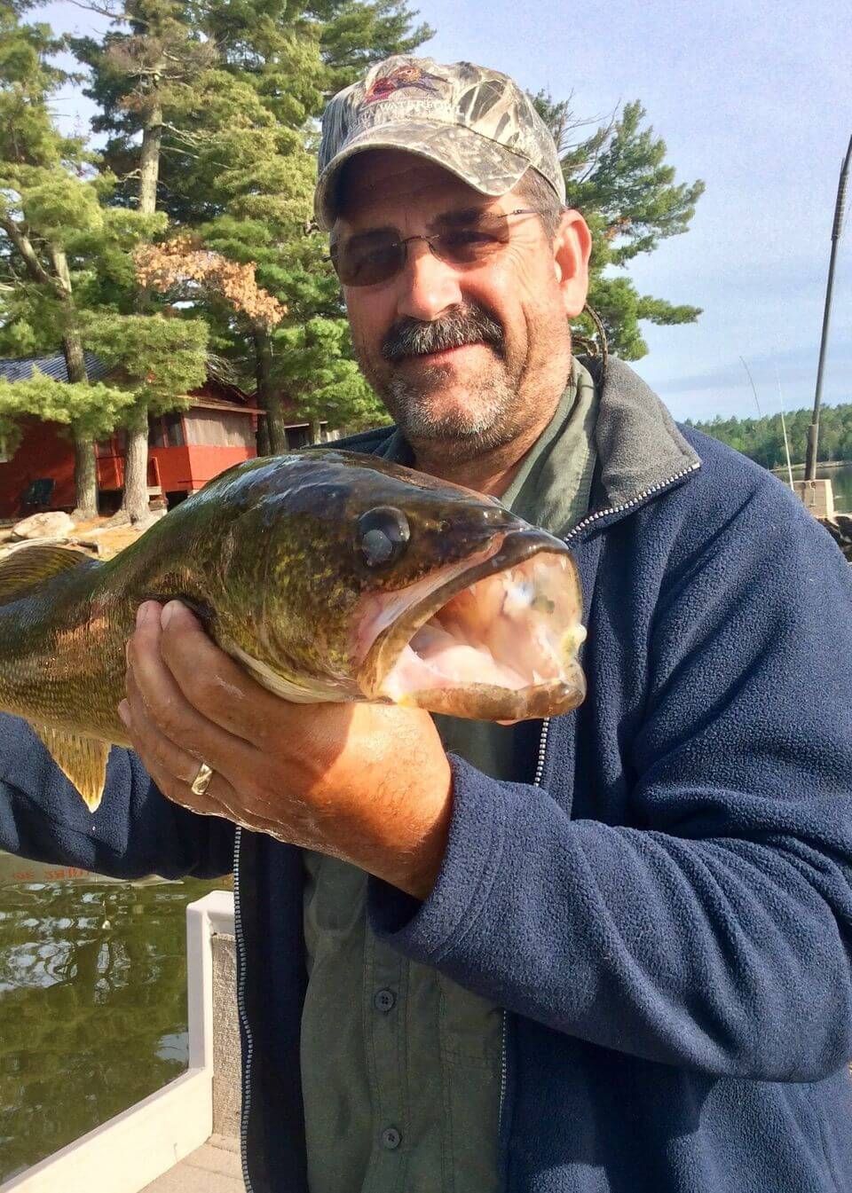 Smiling person holding a large fish by a lake, with trees and a cabin in the background