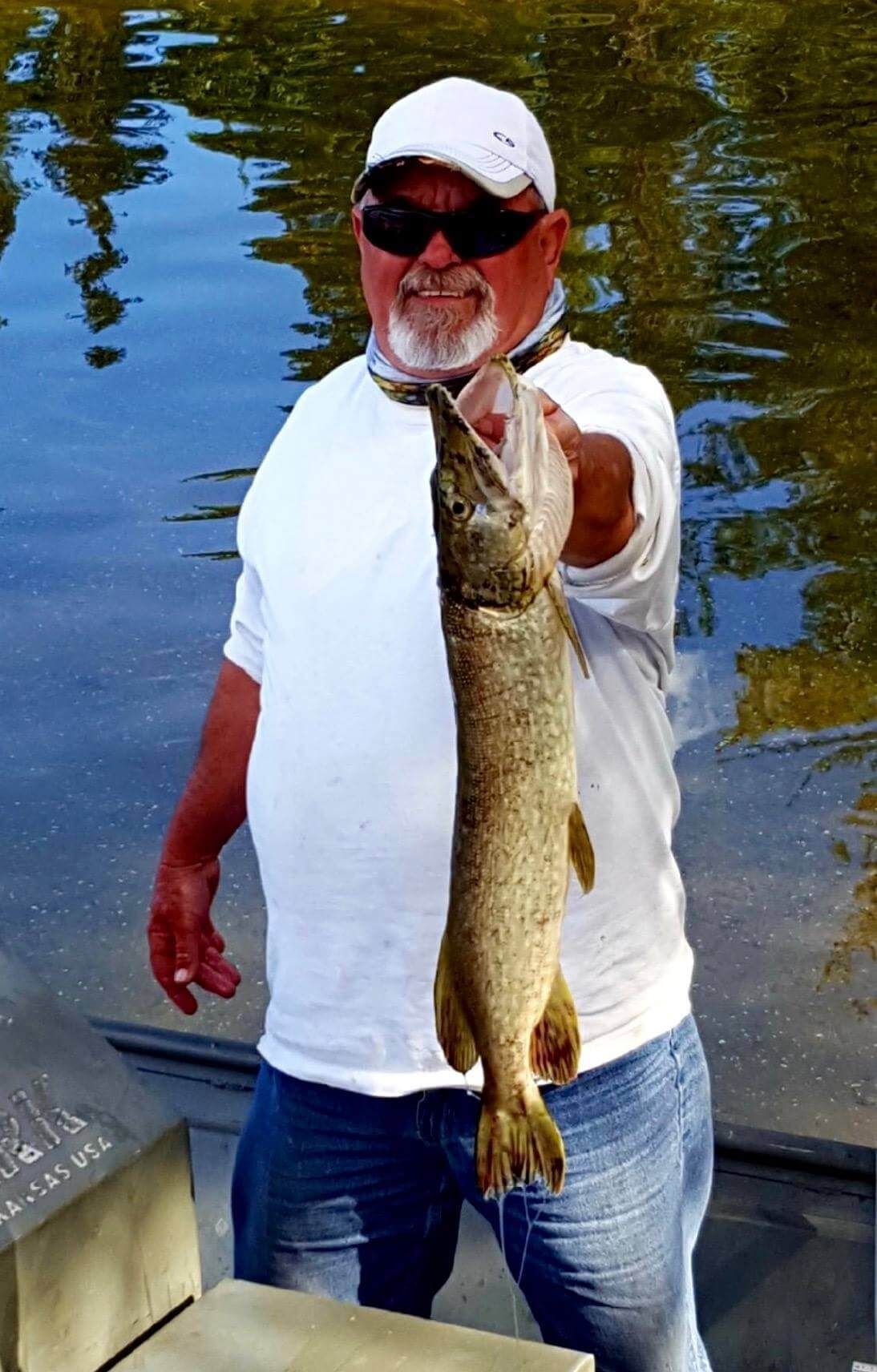 Angler in white shirt and cap holding up a large fish by a calm riverbank