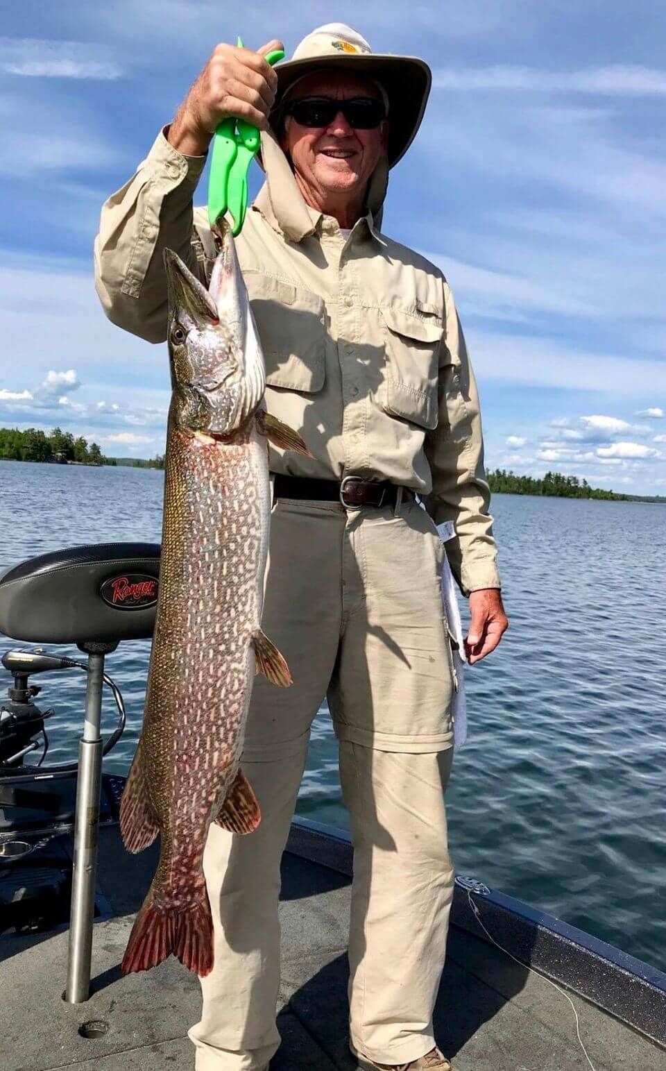 Person on a boat holding a large fish above a lake, wearing a hat and sunglasses.