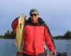 Person in a red jacket holding a large fish by a lake under a blue sky.
