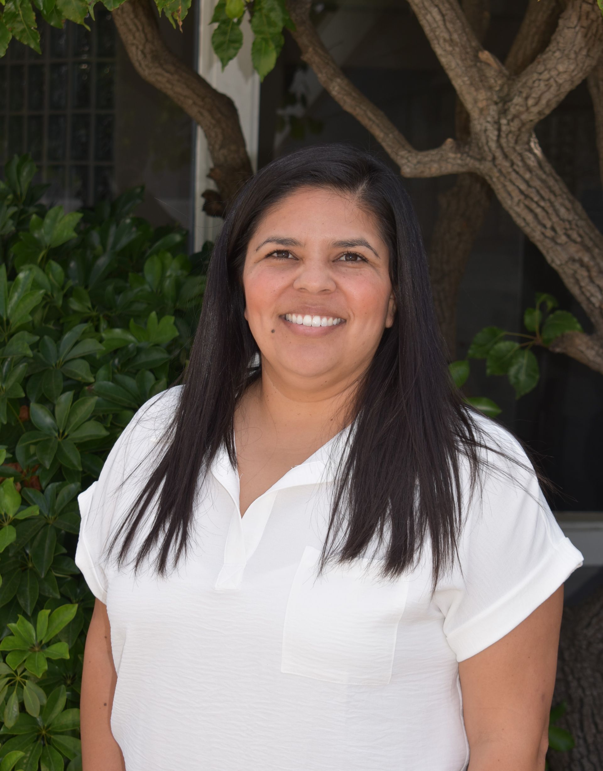 A woman in a white shirt is smiling in front of a tree.