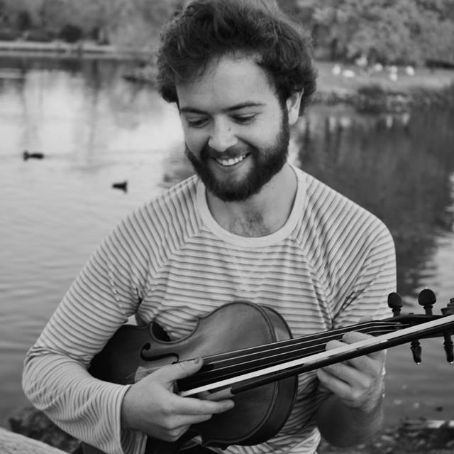 bearded man sitting by a lake, strumming a violin