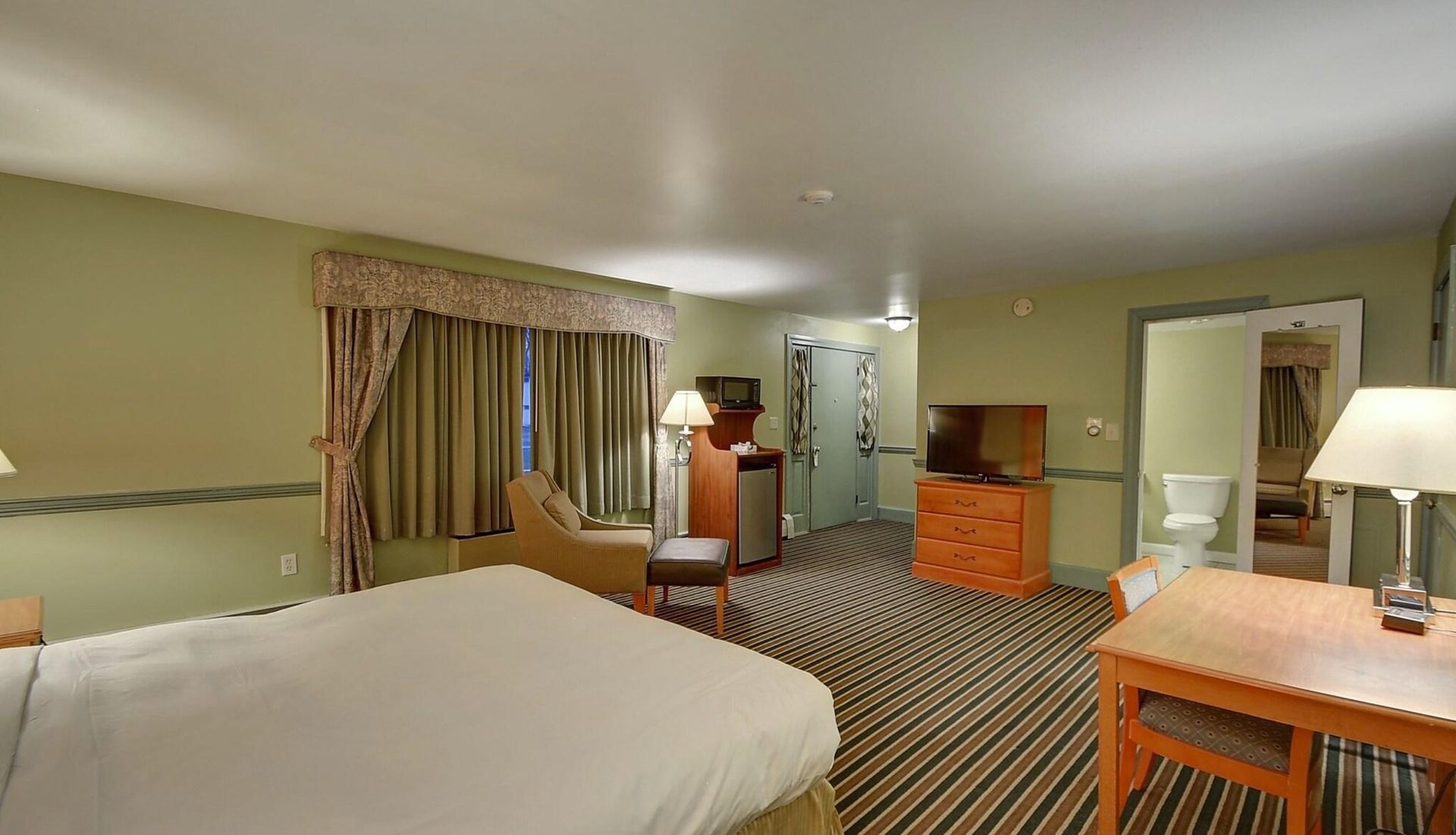 Hotel room with bed, desk, lamps, striped carpet, and curtained windows