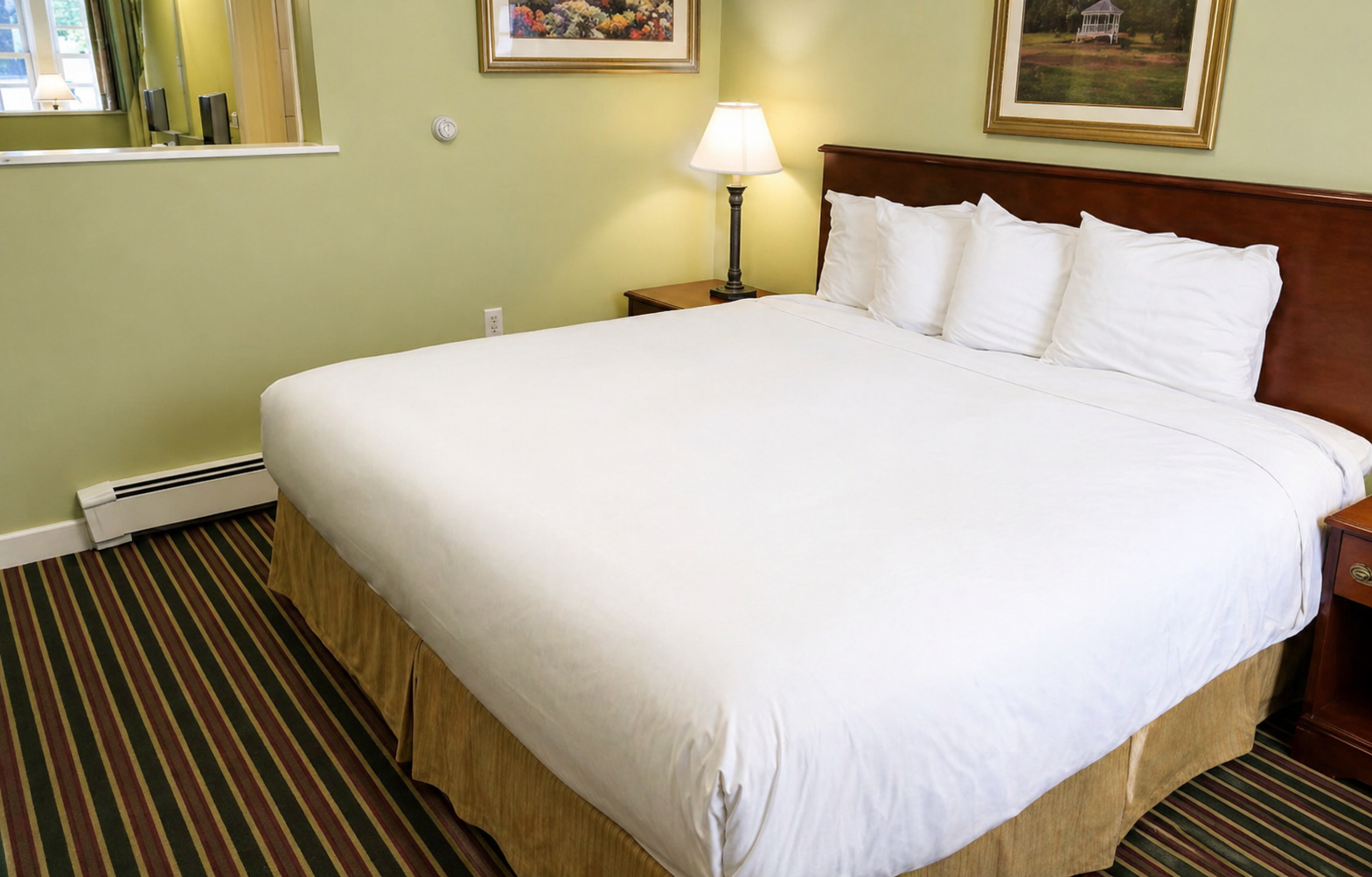 Neatly made hotel bed with white linens, striped carpet, lamp, and framed wall art