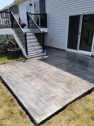 Concrete patio with wood-grain pattern next to house with deck and stairs, surrounded by grass.