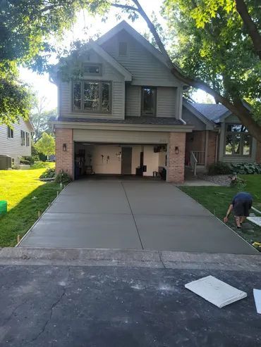 New concrete driveway leading to a two-story house with an open garage. Man works on the side.