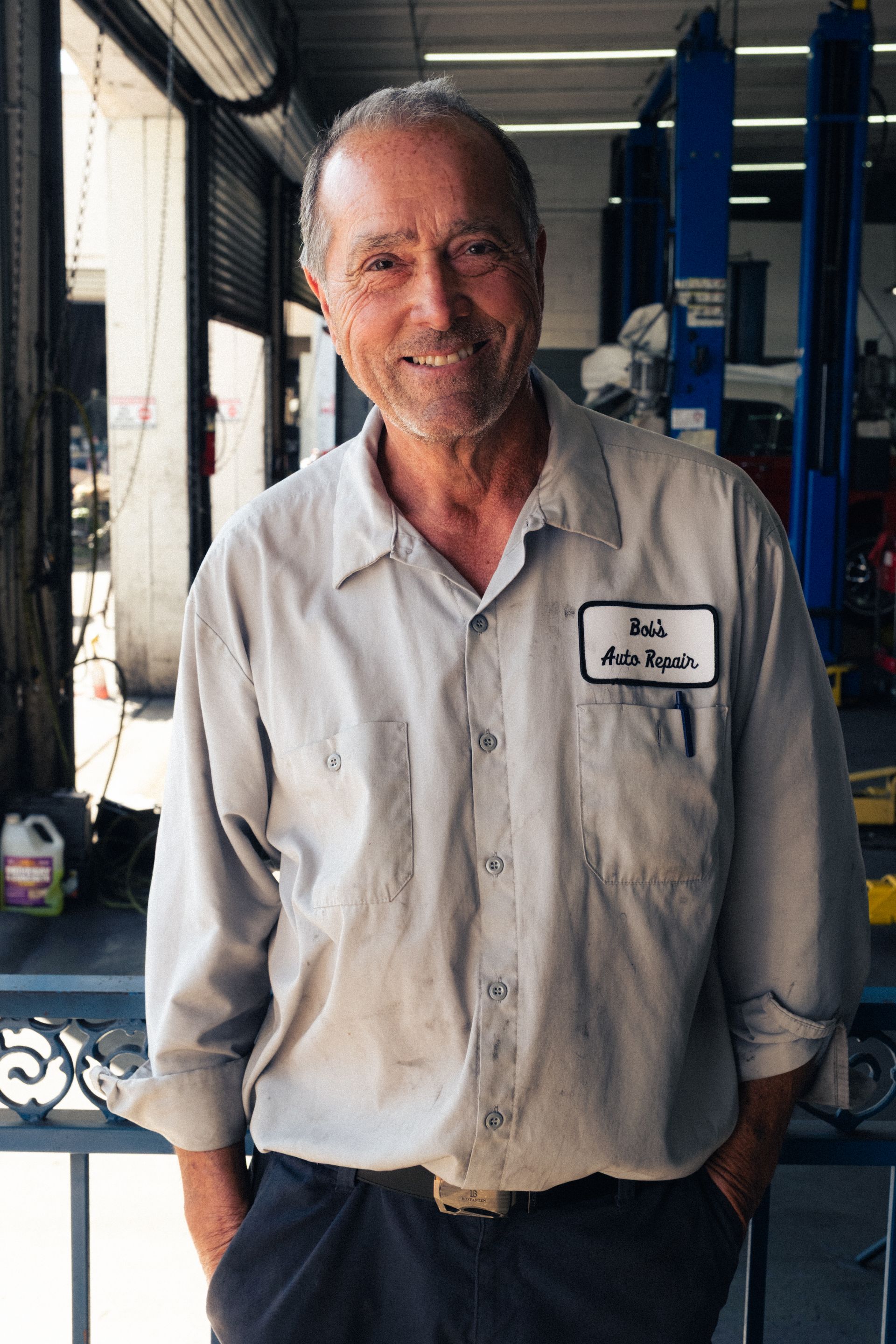 Smiling man in a light work shirt stands in a workshop, hands in pockets, with machinery behind him