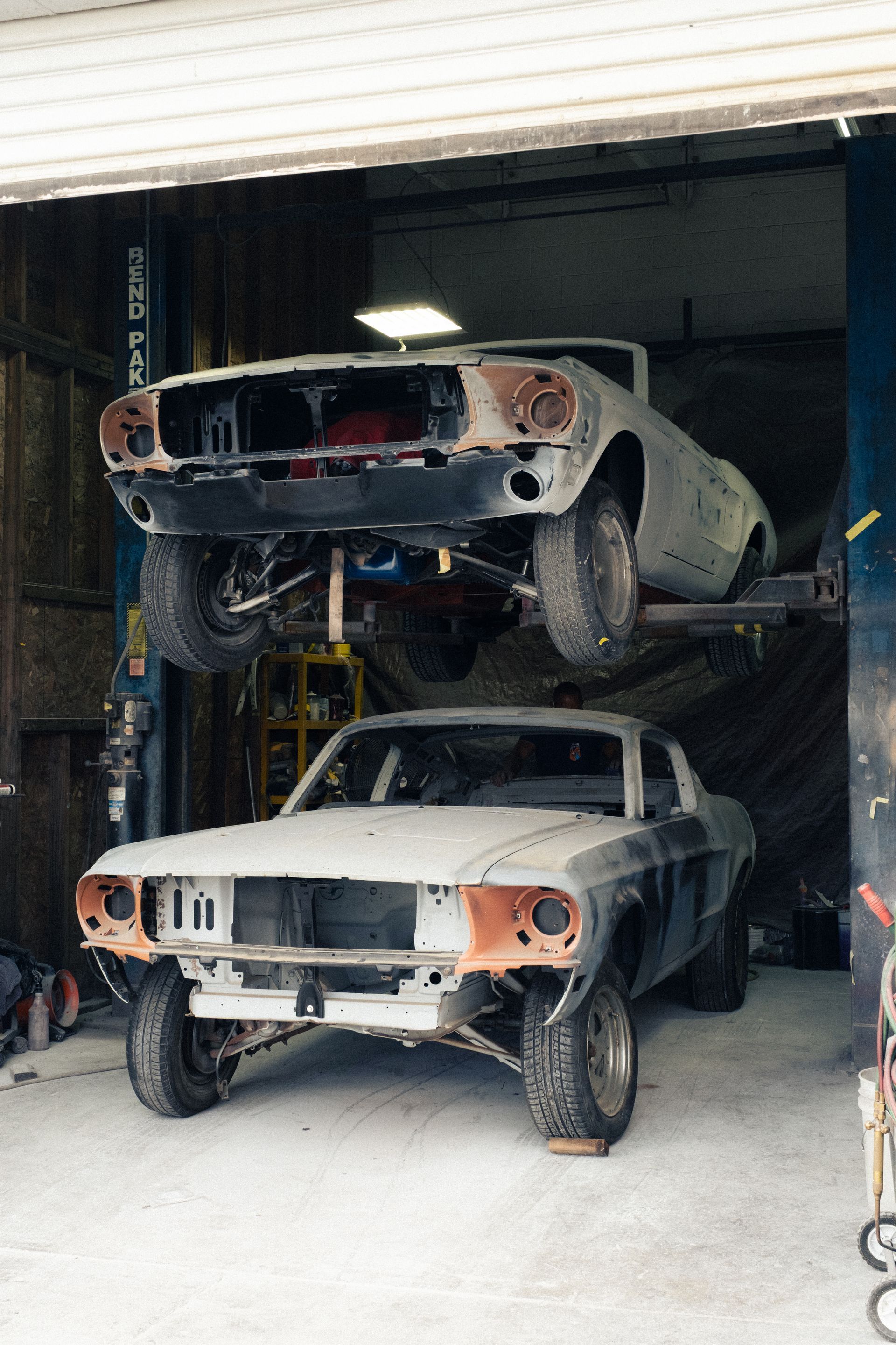 Two vintage cars in a garage, one stacked above the other, both partly restored with missing front parts