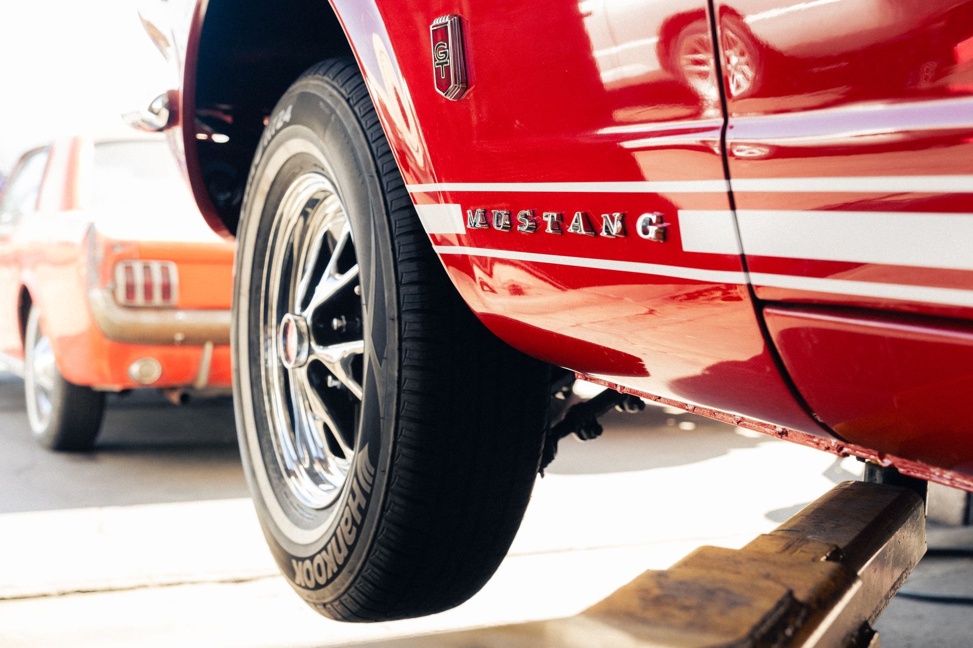 Red Mustang rear wheel lifted on a car jack, with another orange car in the background