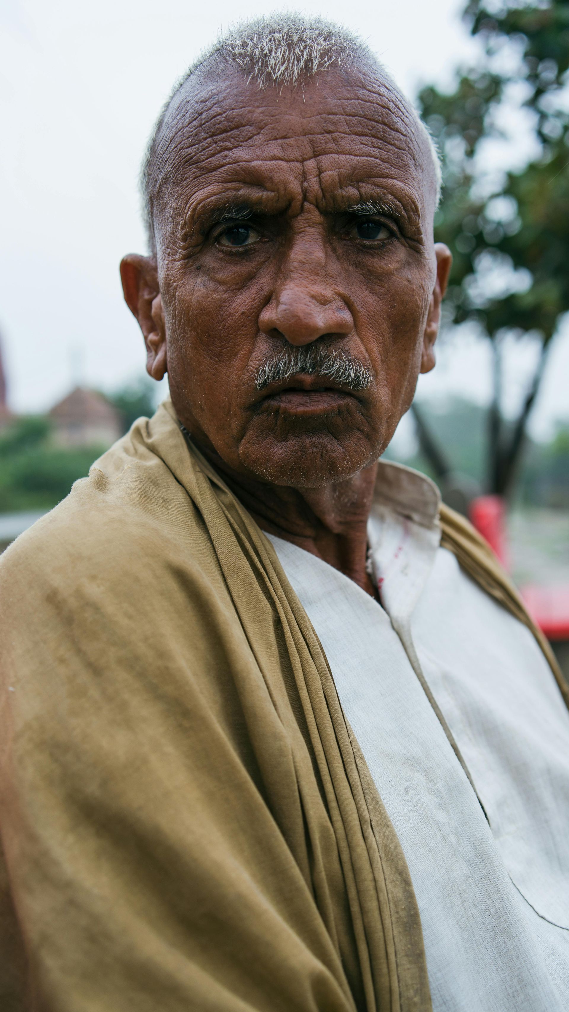 Older man with wrinkled face, mustache, looking forward; wearing white shirt, tan shawl.