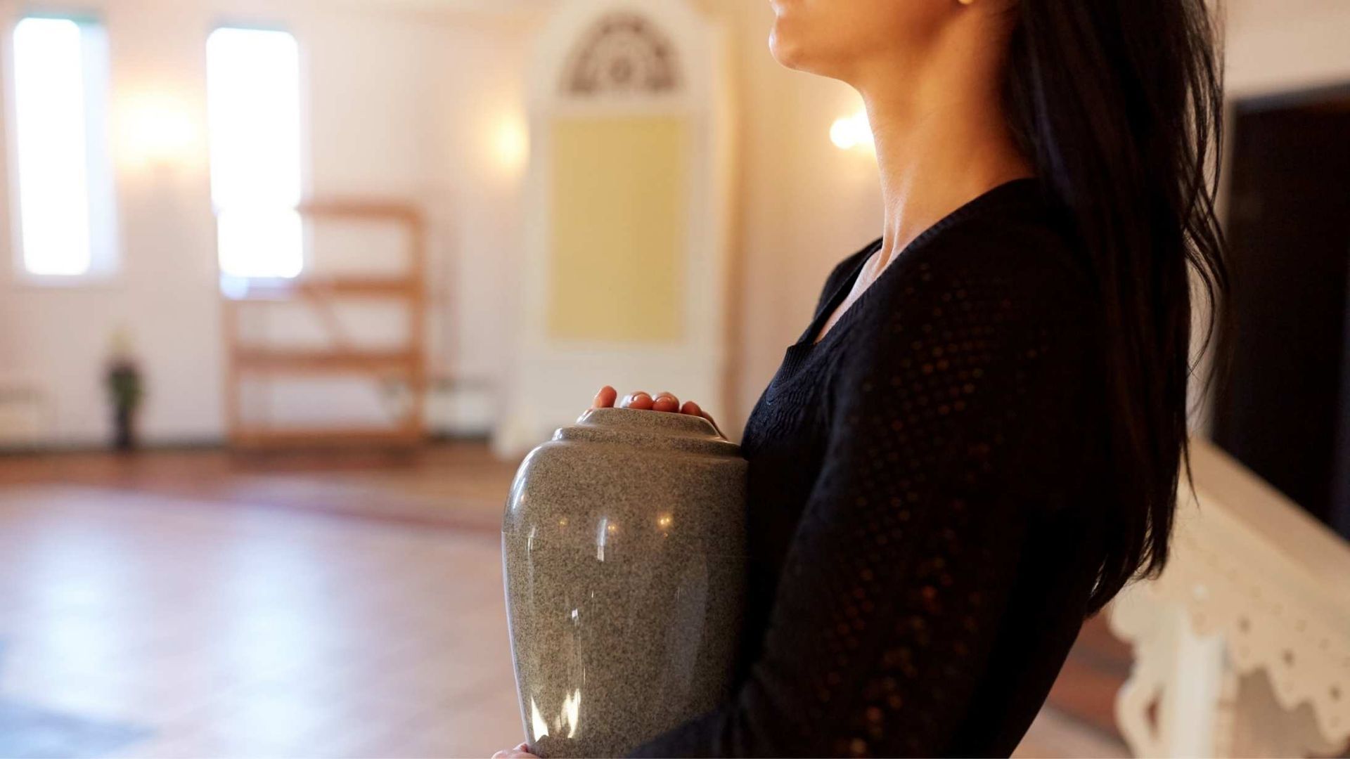 Woman holding a gray urn in a light-filled room, possibly a memorial or funeral setting.