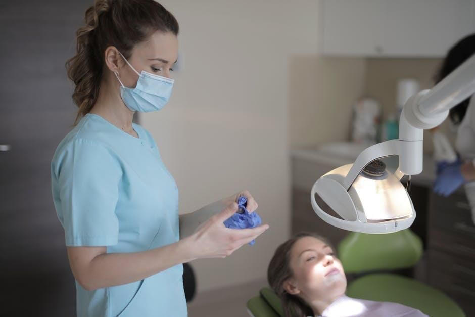 A dental professional in blue scrubs prepares a blue glove next to a patient reclining in a chair under an exam light.