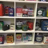 A white shelving unit displaying a collection of vintage oil and antifreeze tins with various colorful brand logos.