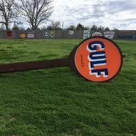 A large, circular orange Gulf Dealer sign lying flat on a grassy field in front of a fence lined with vintage signs.