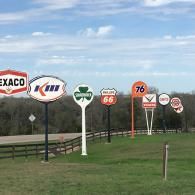 A row of vintage gas station signs standing in a grassy field against a blue, cloudy sky.