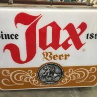 A vintage-style white and tan sign for Jax Beer, featuring large red lettering and a circular illustration of a rider.