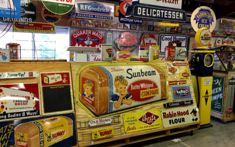 A wide collection of vintage advertising signs and a yellow gas pump displayed inside a rustic shop setting.