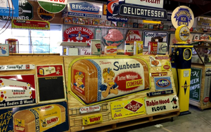 A wide collection of vintage advertising signs and a yellow gas pump displayed inside a rustic shop setting.