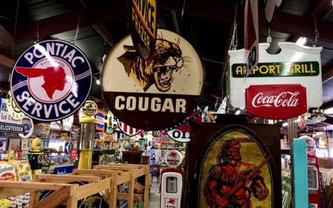A collection of vintage signs, including Pontiac Service and Cougar logos, hangs from the ceiling in a shop filled with items.