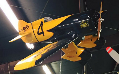 A black and yellow Gee Bee Model Z race plane suspended from the ceiling of an indoor hangar.