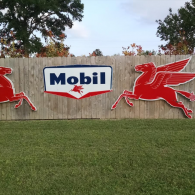 A wooden fence displays a blue and white Mobil sign flanked by two large, red flying horse logos on a grassy lawn.