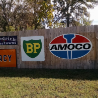 Vintage fuel and tire company signs mounted on a wooden fence outdoors above a grassy lawn.