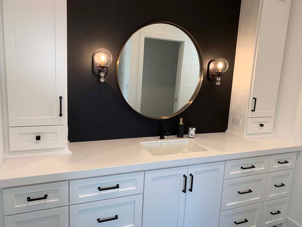 A bathroom with white cabinets , drawers , a sink and a round mirror.