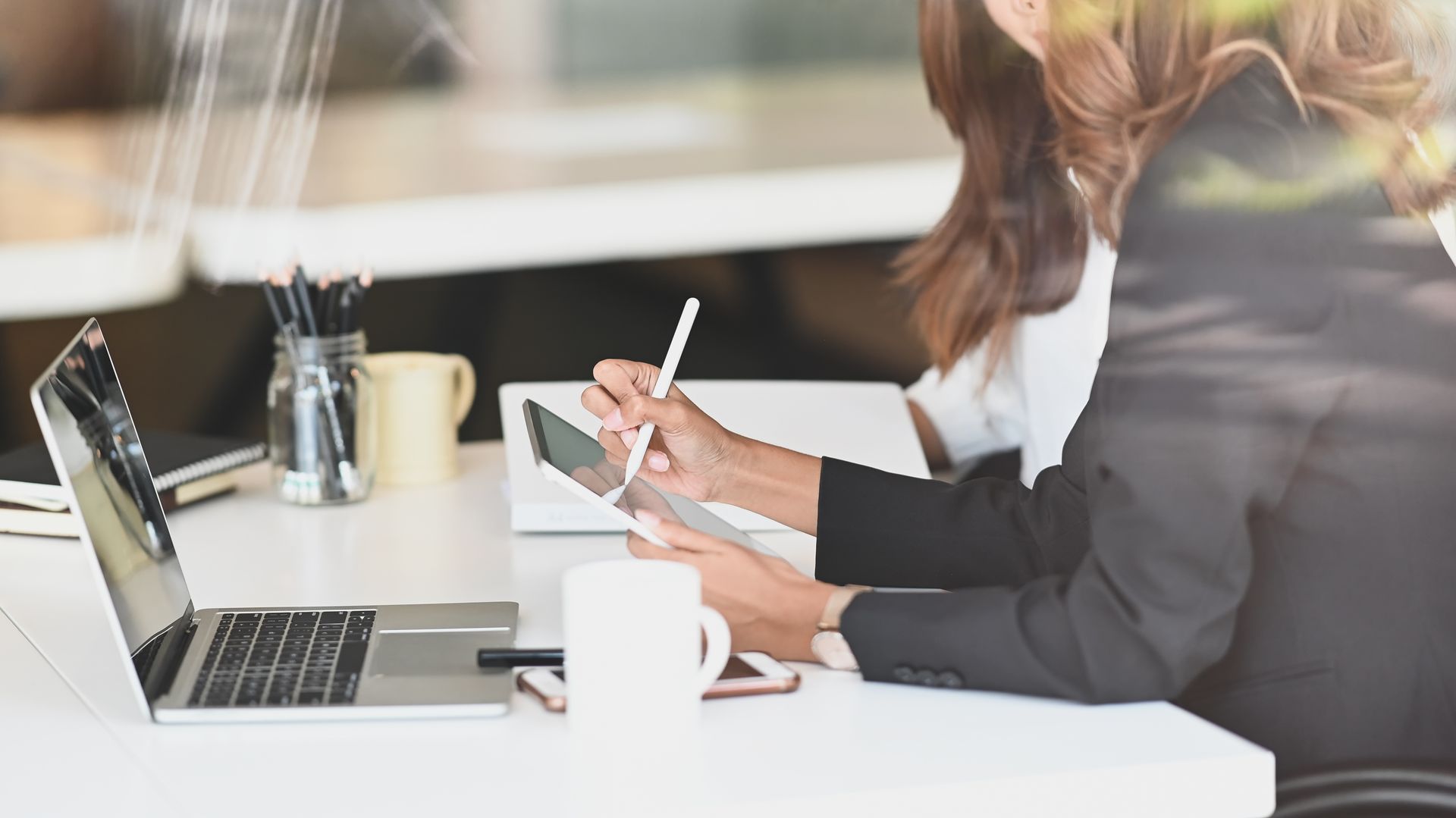 Two people in business attire working at a desk with a laptop and tablet, white mug, natural light.