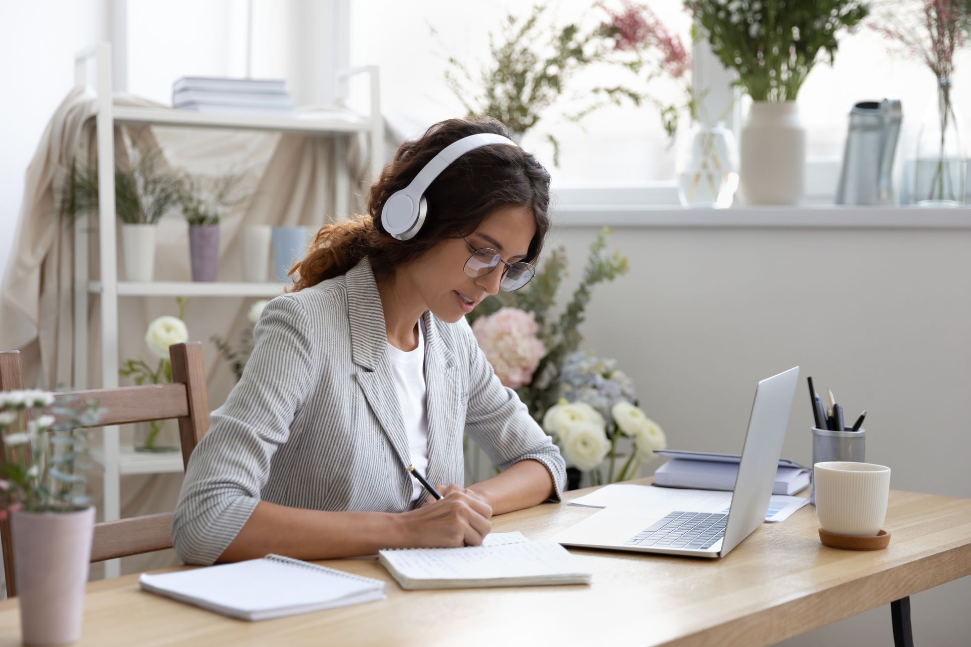 Woman wearing headphones and glasses, writing in a notebook at a desk with a laptop, flowers, and a window.