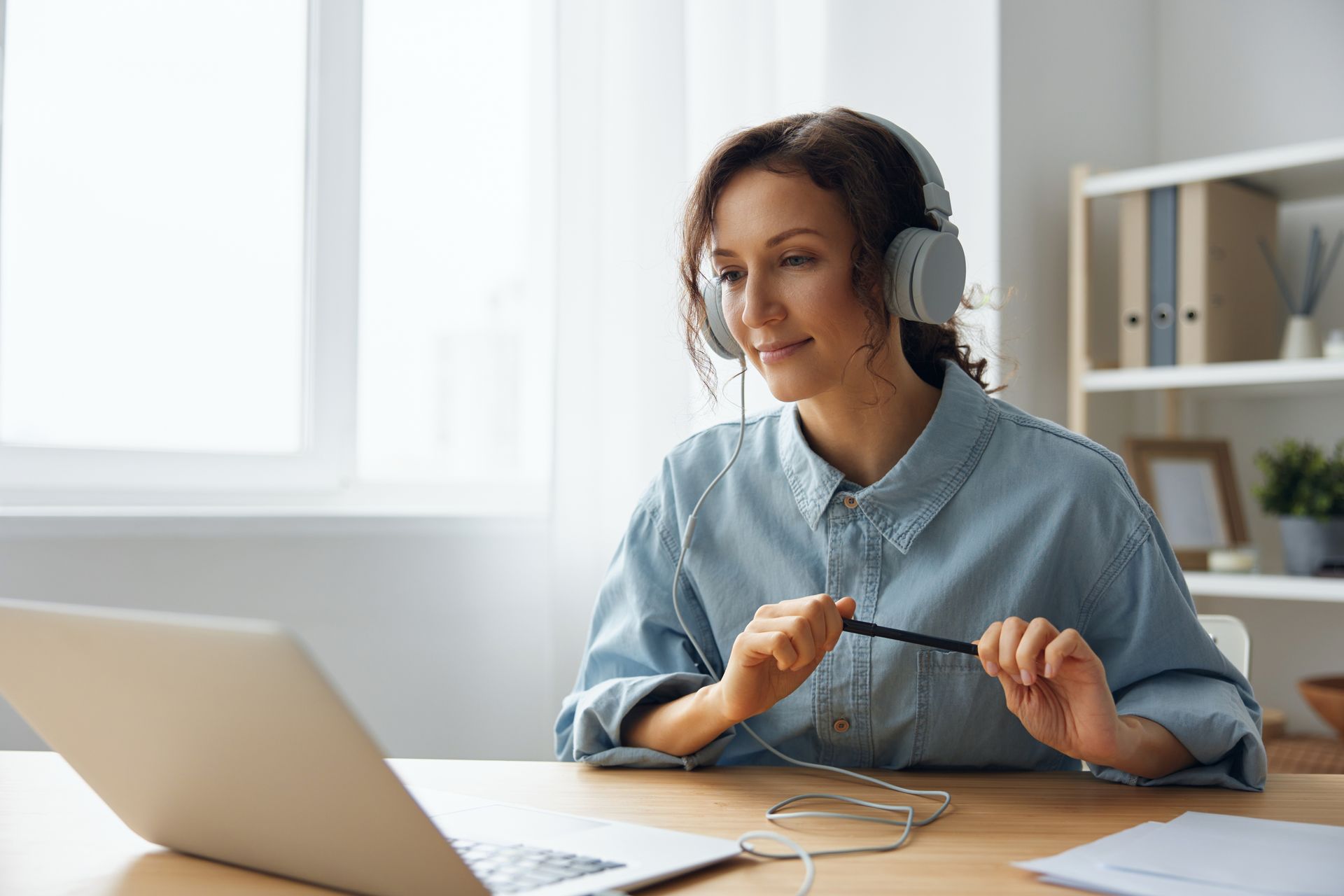 Woman in headphones, at desk, looking at laptop screen, holding pen, light blue shirt, indoors.