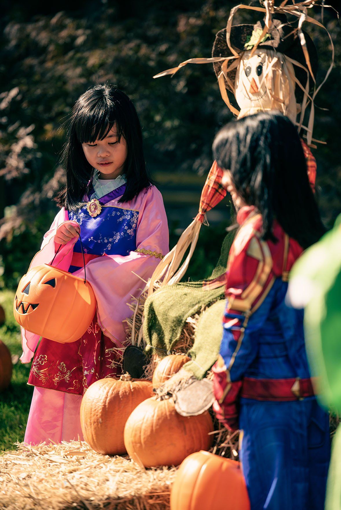 Two girls in costumes are standing next to pumpkins and a scarecrow.