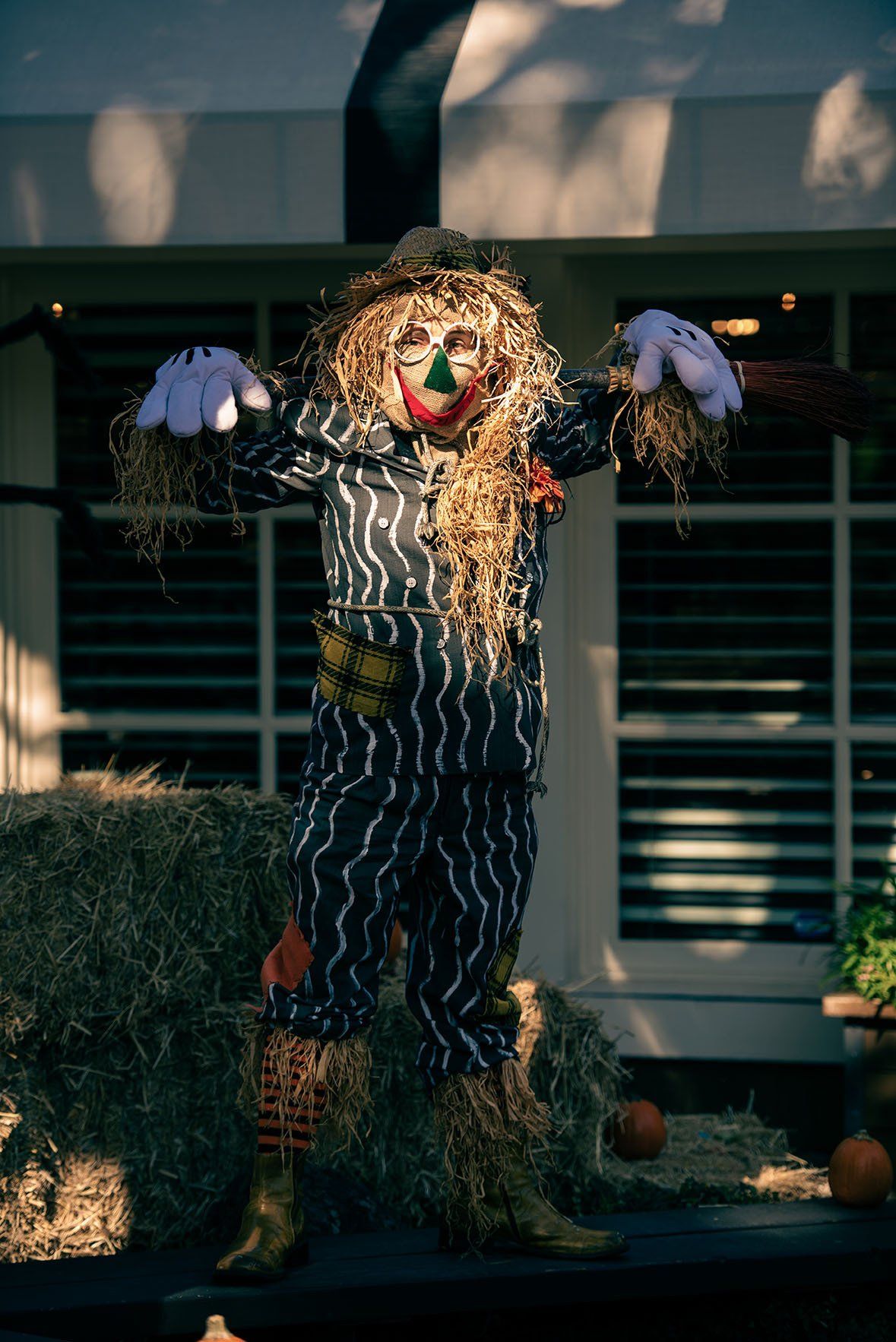 A scarecrow is standing in front of a house holding hay bales.